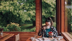 A person sitting at a wooden table with a cup, surrounded by plates and utensils, in a room with large windows and a view of greenery.