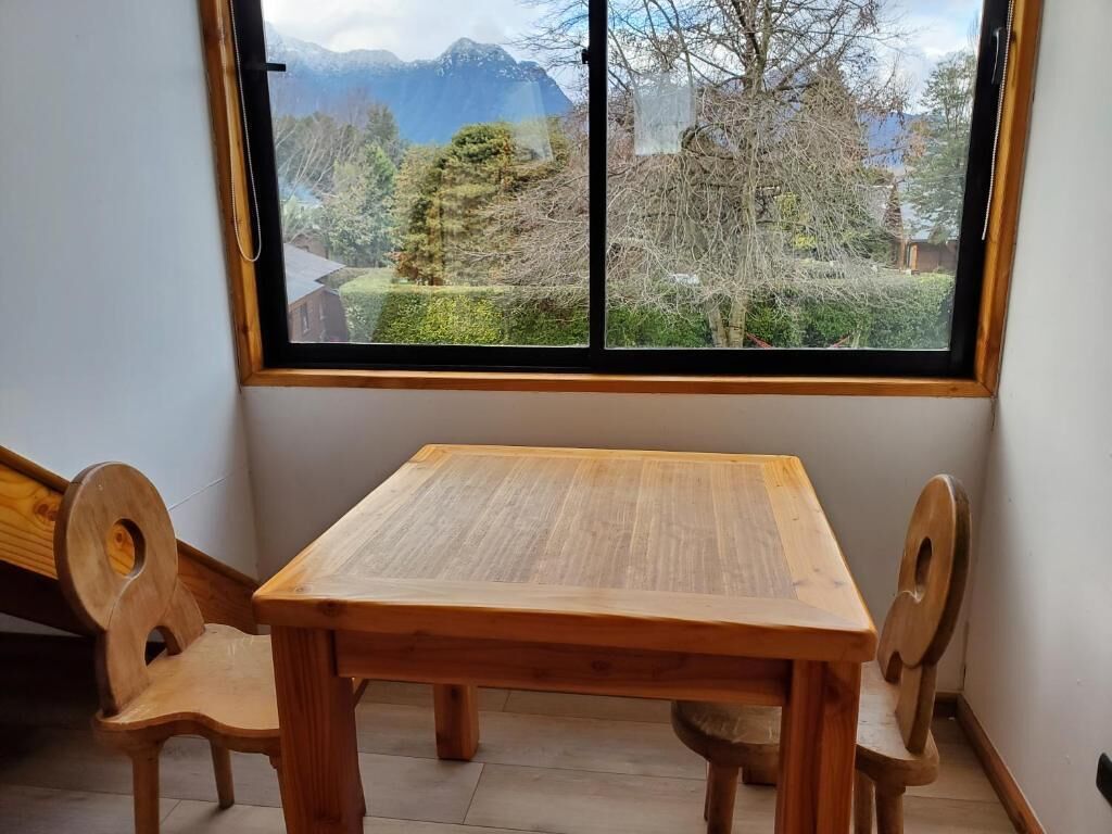 Una mesa de madera con dos sillas en una habitación con un ventanal grande que da a un jardín y montañas.