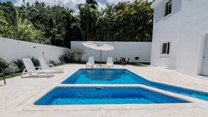 A rectangular swimming pool with two lounge chairs and an umbrella, surrounded by a white wall and lush greenery. - Villa Belle Mar with a pool and within walking distance of the beach!  (Puerto Plata)