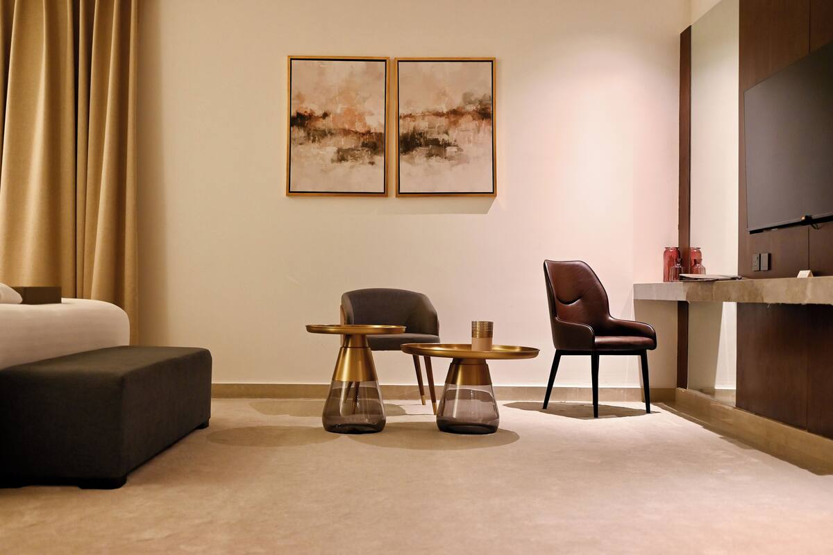 A modern hotel room with a bed, two side tables, a chair, and a desk. There are two framed artworks on the wall.