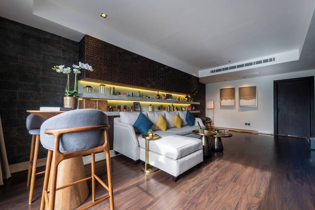 A modern living room with a white sofa, blue and yellow cushions, wooden chairs, a coffee table, and a decorative shelf with various items.