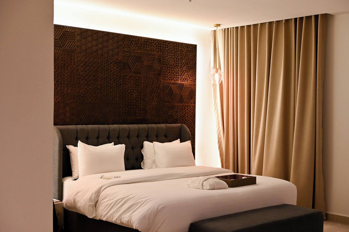 A modern hotel room with a tufted headboard, a large bed with white linens, a bedside table, and a large window with beige curtains.