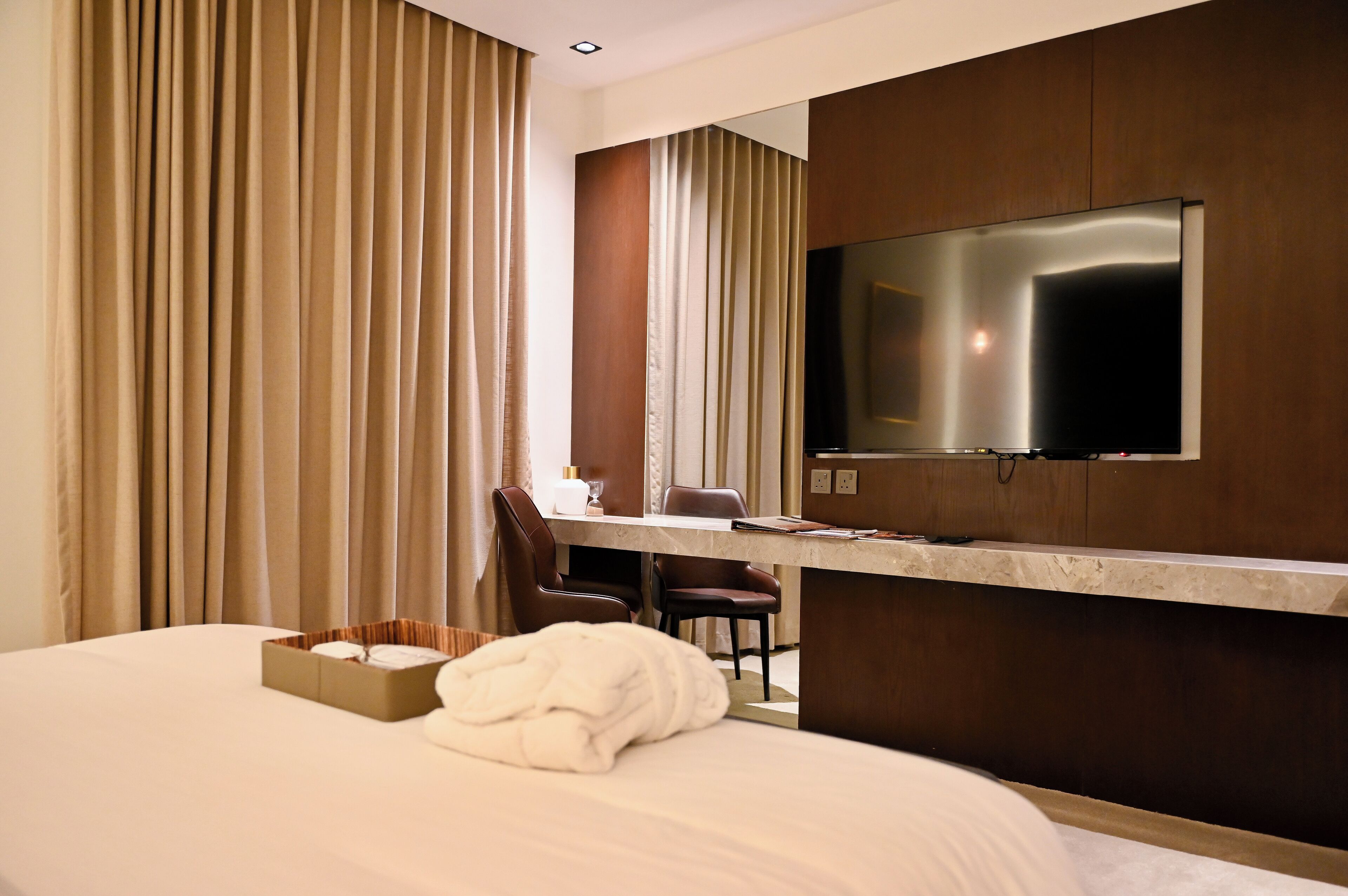 A hotel room with a large bed, a desk with a chair, a TV mounted on the wall, and a window with curtains.