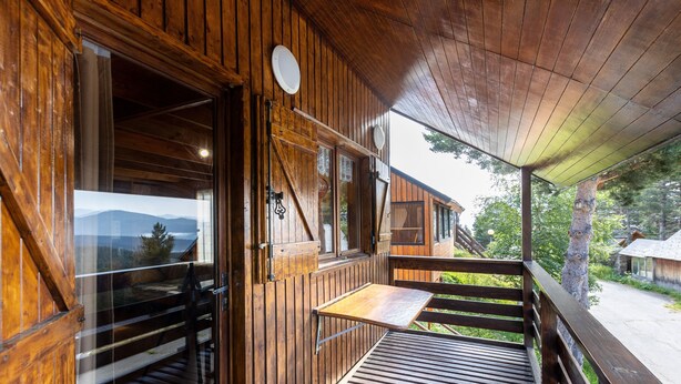 Una cabaña de madera con terraza de madera, una ventana con vista a las montañas y una barandilla de madera.