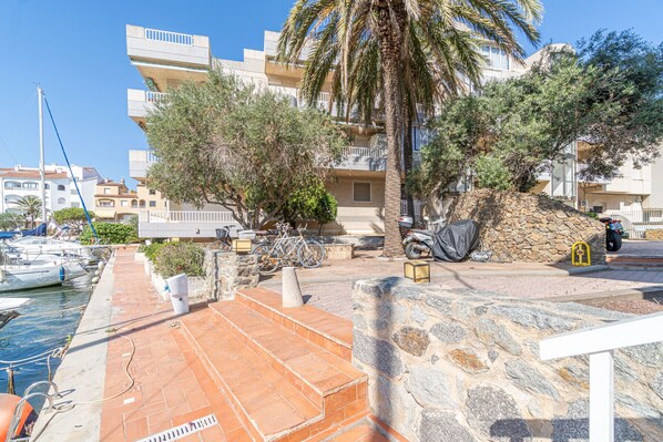 A coastal building with a stone staircase leading to a terrace, surrounded by palm trees and a marina with boats.