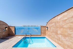 A rooftop swimming pool with clear blue water, surrounded by beige tiled walls and a wooden deck.