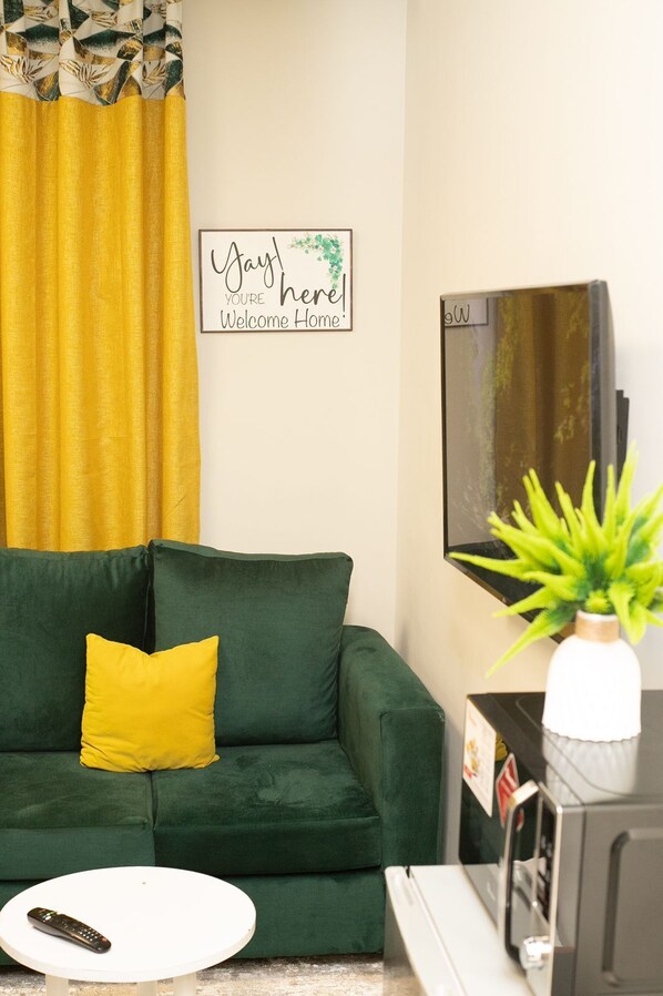 A living room with a green sofa, a yellow pillow, a framed welcome sign, a television, and a plant.