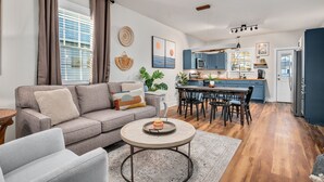 A modern living room with a grey sofa, a round coffee table, and a dining area with a wooden table and chairs. A kitchen with blue cabinets and a white refrigerator is visible in the background.
