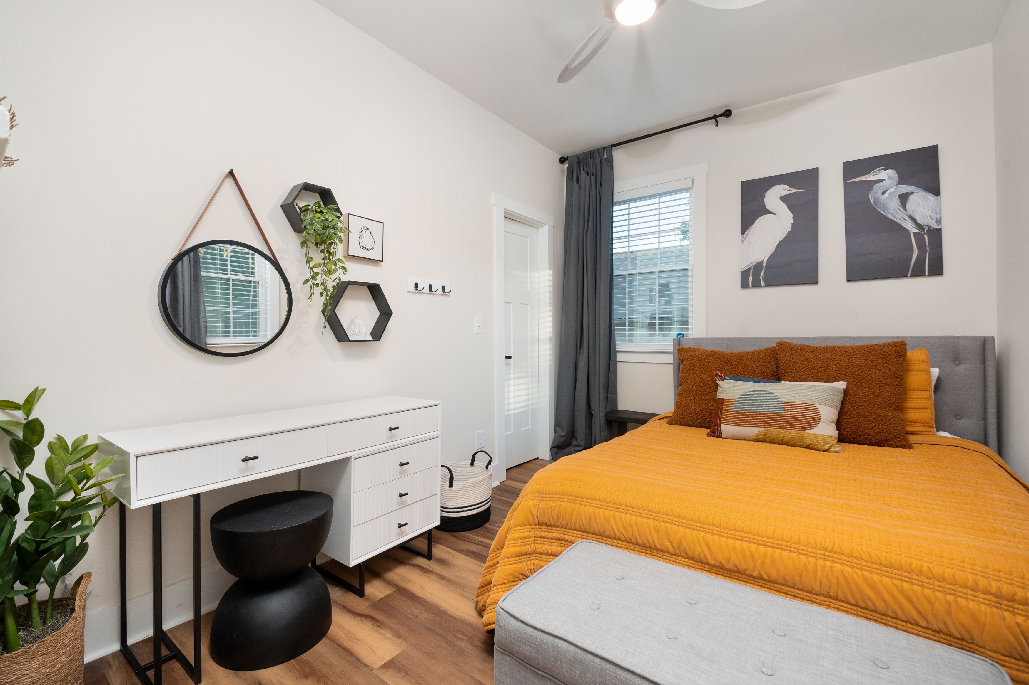 Un dormitorio moderno con un escritorio blanco, una cama grande con colcha color mostaza y dos cuadros de pájaros colgados en la pared.