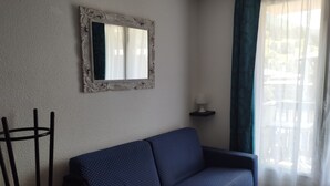 A living room with a blue sofa, a mirror on the wall, a lamp on a side table, and a window with curtains. - Studio 4 Personnes Avec Balcon aux Pieds des Remontées Mécaniques (Allos)
