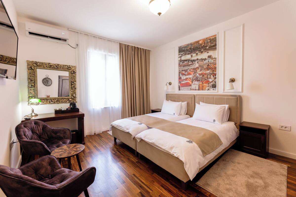 A hotel room with a large bed, two bedside tables, a desk with a chair, and a large window with curtains.