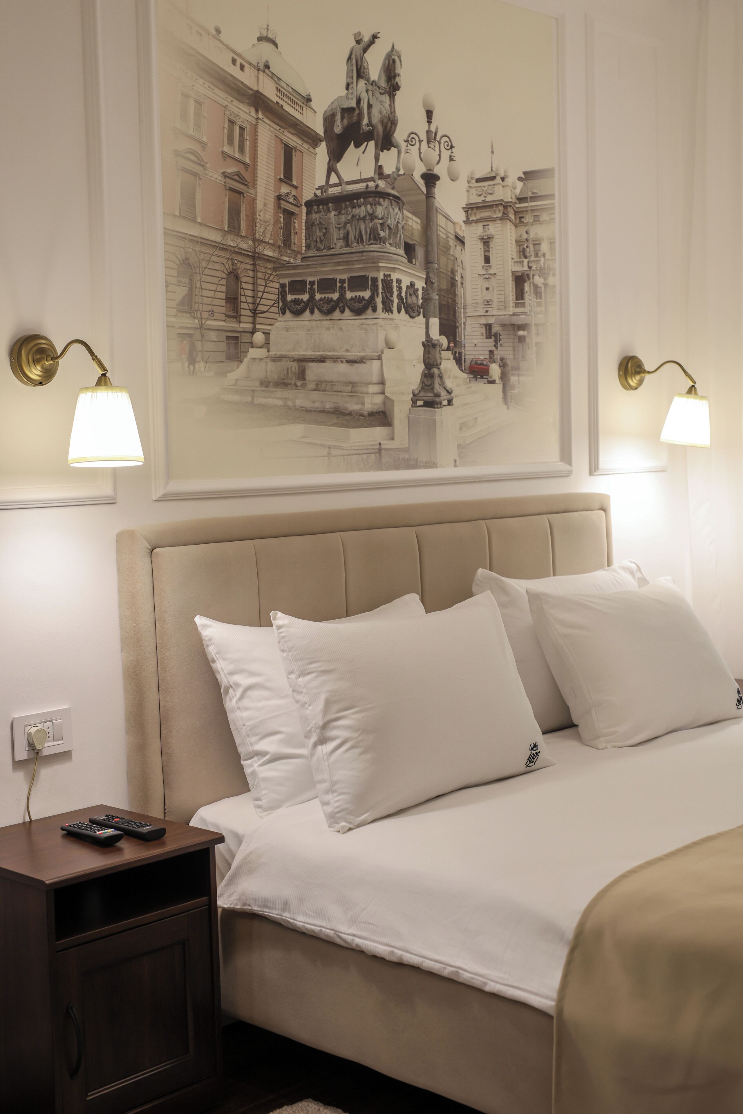 A hotel room with a large bed, a nightstand, a lamp, and a wall-mounted painting of a fountain.