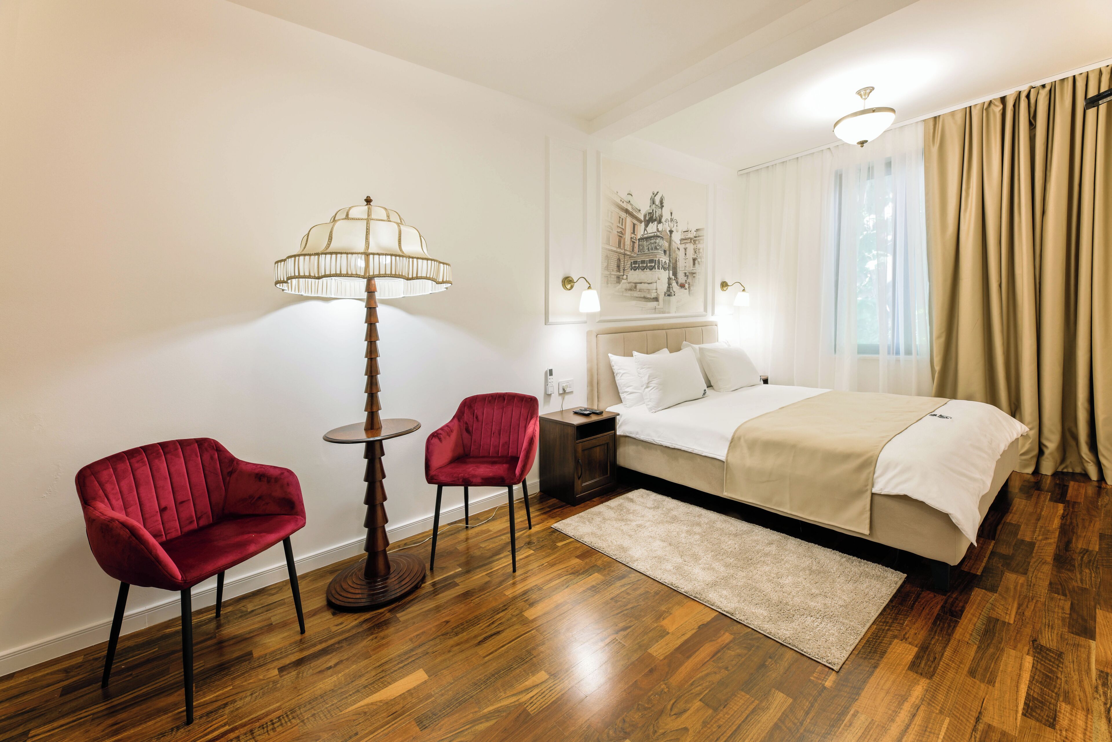 Habitación de hotel con una cama, dos sillas rojas, una lámpara de pie, una mesita de noche y un ventanal con cortinas.