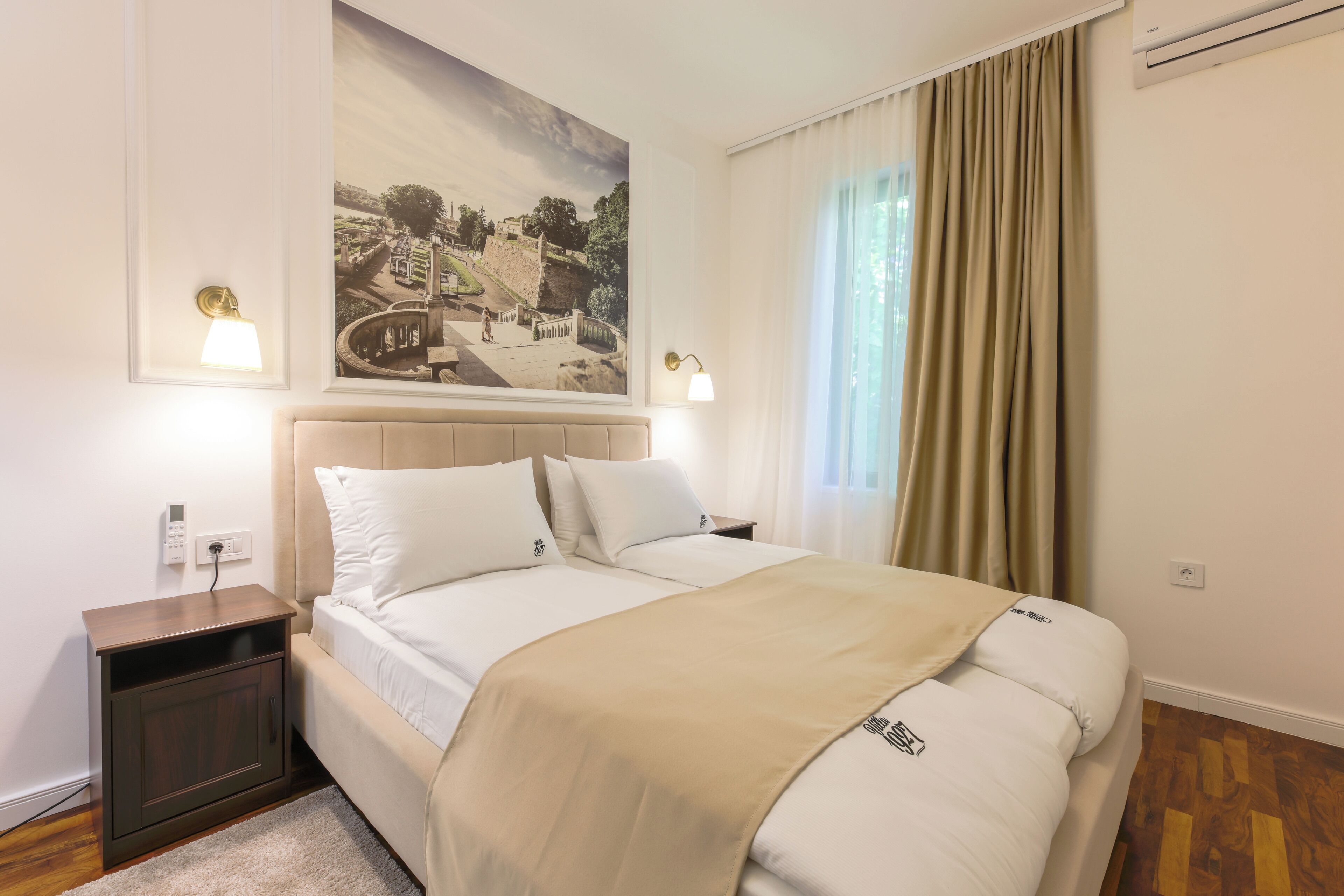Una habitación de hotel ordenada con una cama grande, mesitas de noche, un cuadro colgado en la pared y una ventana con cortinas.