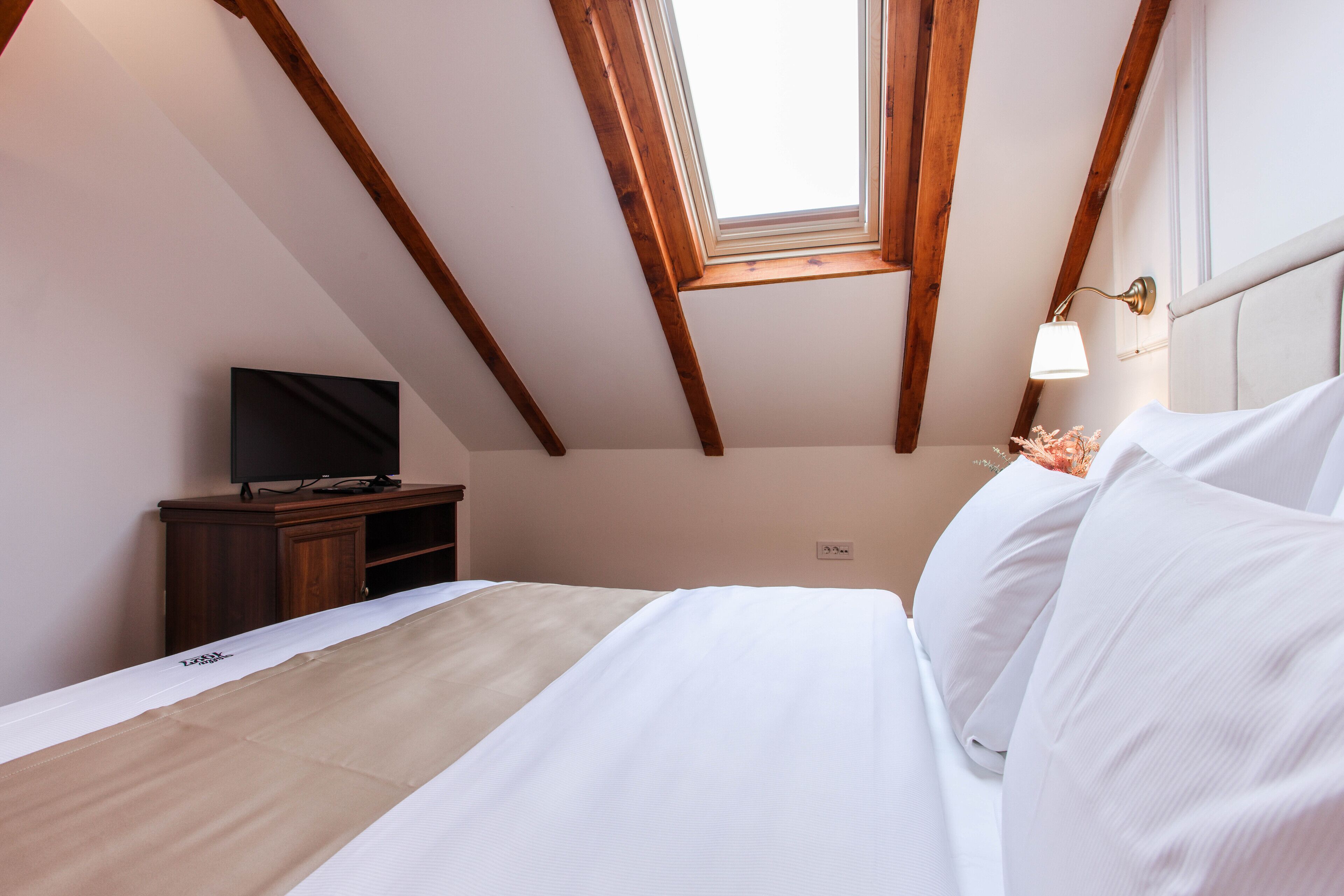 A bedroom with a bed, a TV on a wooden cabinet, a skylight, and a wall-mounted lamp.