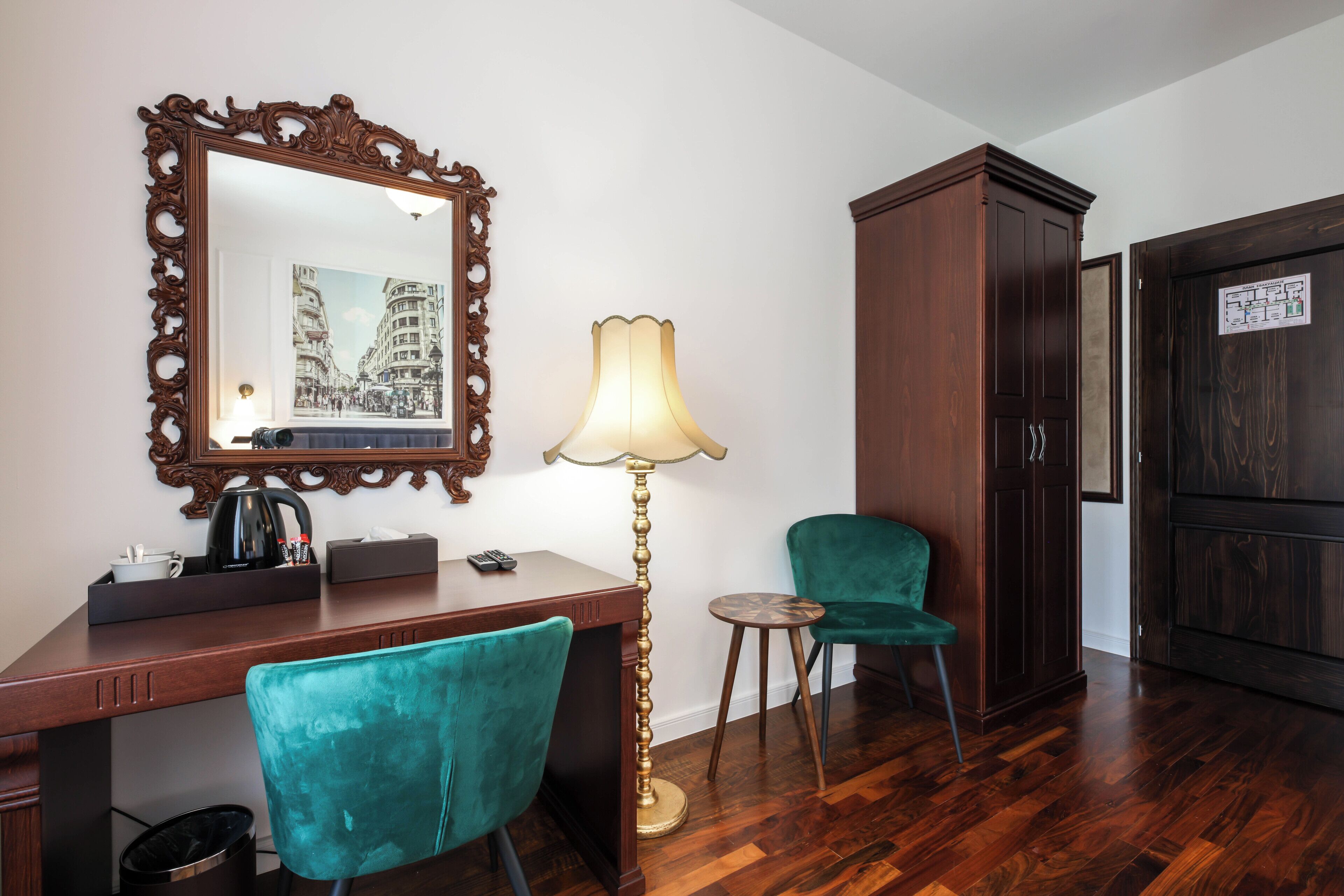 A hotel room with a wooden desk, a teal chair, a floor lamp, a large mirror, and a wardrobe.