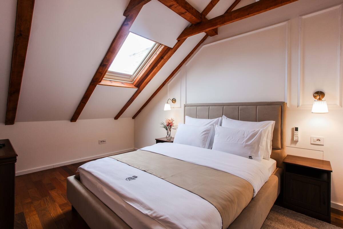 A bedroom with a sloped ceiling, a bed with white bedding, a nightstand with a lamp, and a small table with a vase of flowers.