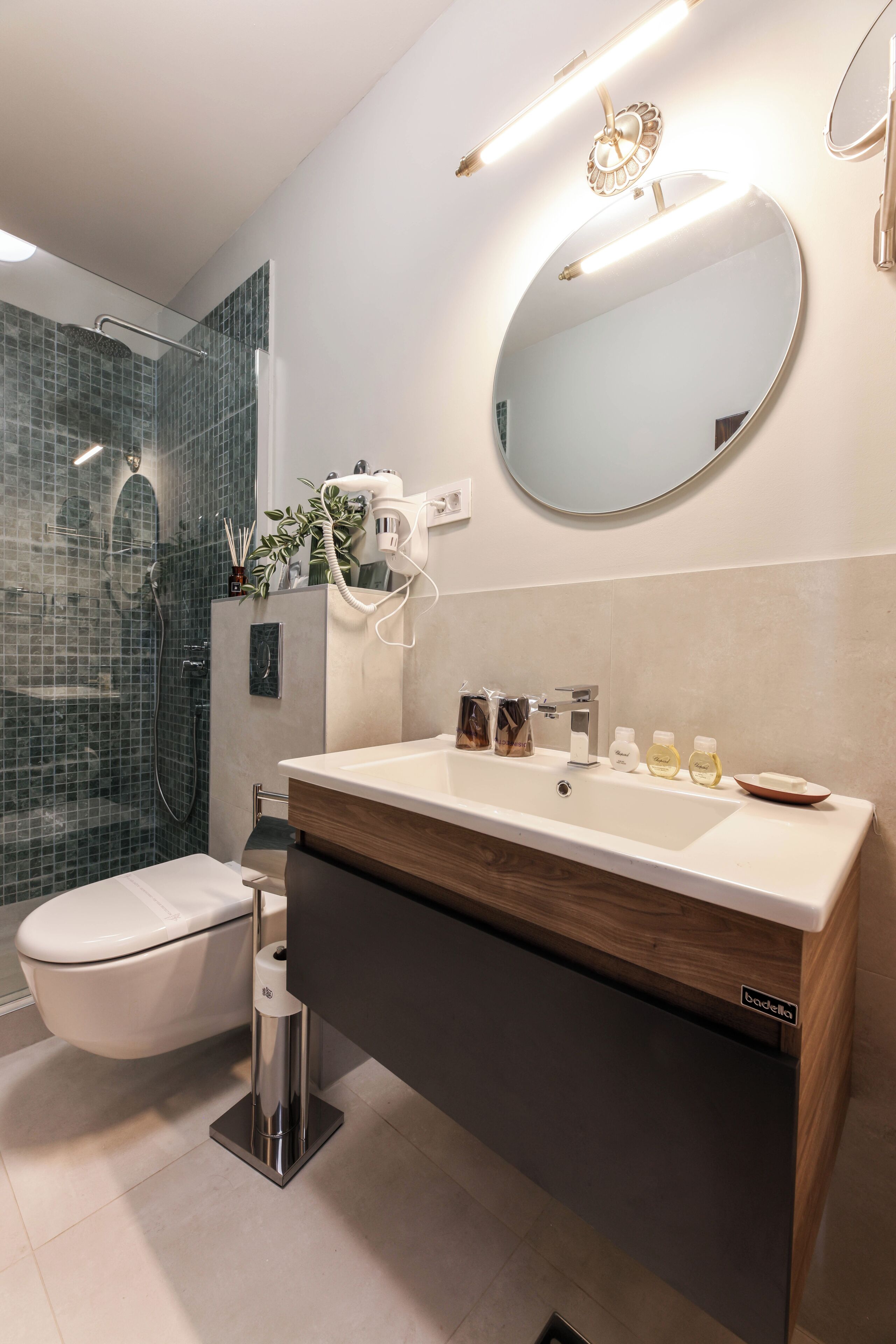 A modern bathroom with a glass shower enclosure, a white toilet, a double sink vanity, and a round mirror.