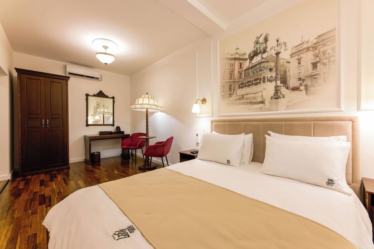 A hotel room with a large bed, a desk with a chair, a mirror, and a wall-mounted lamp.