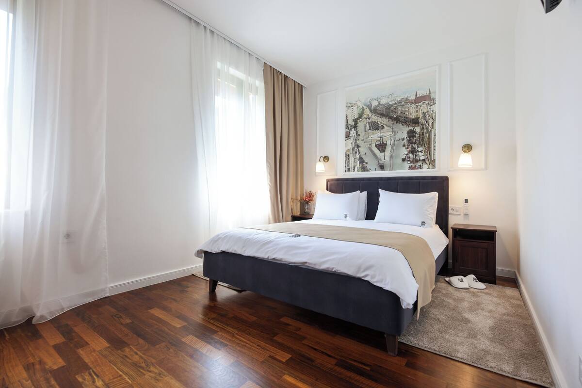 A neatly arranged bedroom with a large bed, a nightstand with a lamp, and a cityscape painting on the wall.