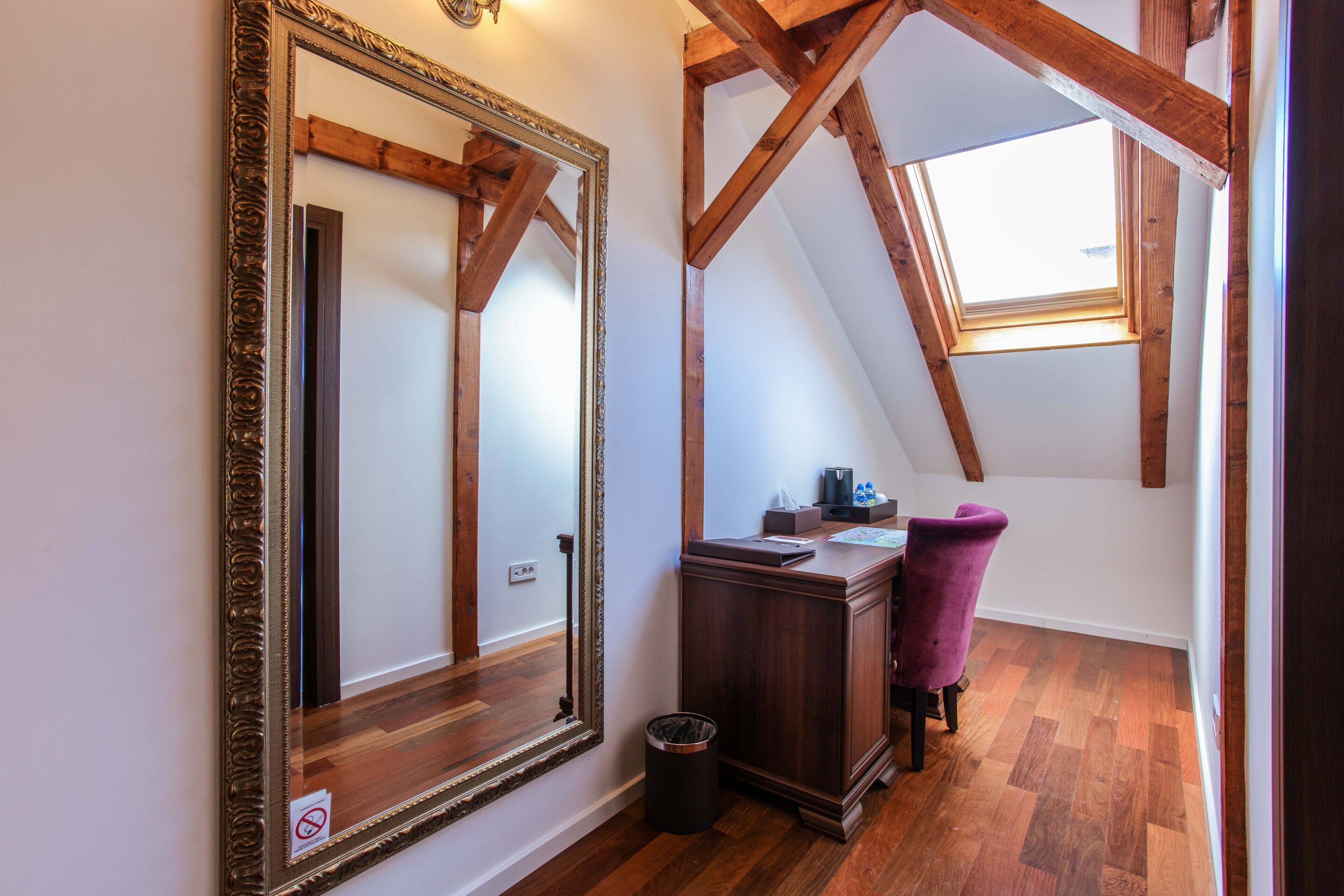 Una habitación con un espejo grande y ornamentado, vigas de madera, un escritorio con una silla y un tragaluz.