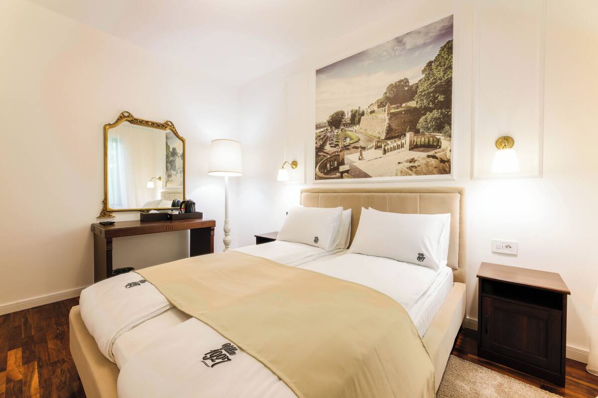 A hotel room with a large bed, a nightstand, a mirror, a lamp, and a painting on the wall.