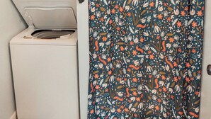 Pieni ja käytännöllinen pyykinhuoltotila, jossa on pesukone ja kuivausrumpu, kukkakuviollinen suihkuverho ja tumma matto lattialla.
