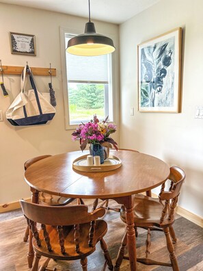 A round wooden dining table with chairs, a hanging light fixture, a window with blinds, and a framed welcome sign. - Lake Top Cottage - Midcoast Maine - New Reno - High-Speed WiFi (Jefferson)