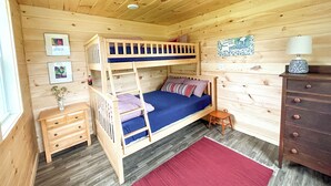 Een houten slaapkamer met een stapelbed, een klein houten nachtkastje, een dressoir en een rode vloerkleed.
