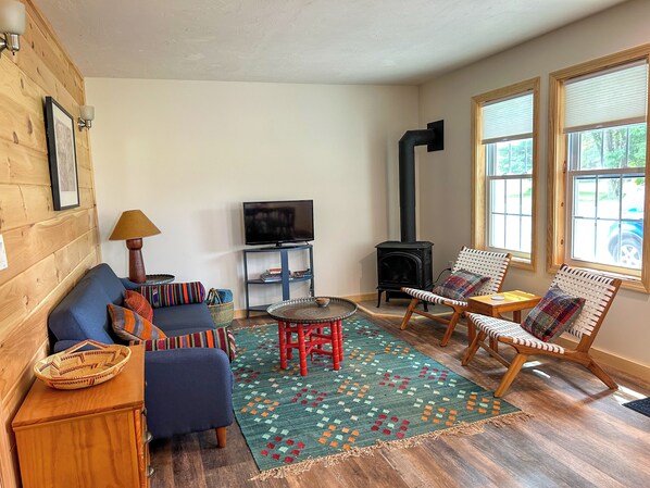 A cozy living room with a sofa, chairs, a TV, a fireplace, and a large window. - Lake Top Cottage - Midcoast Maine - New Reno - High-Speed WiFi (Jefferson)