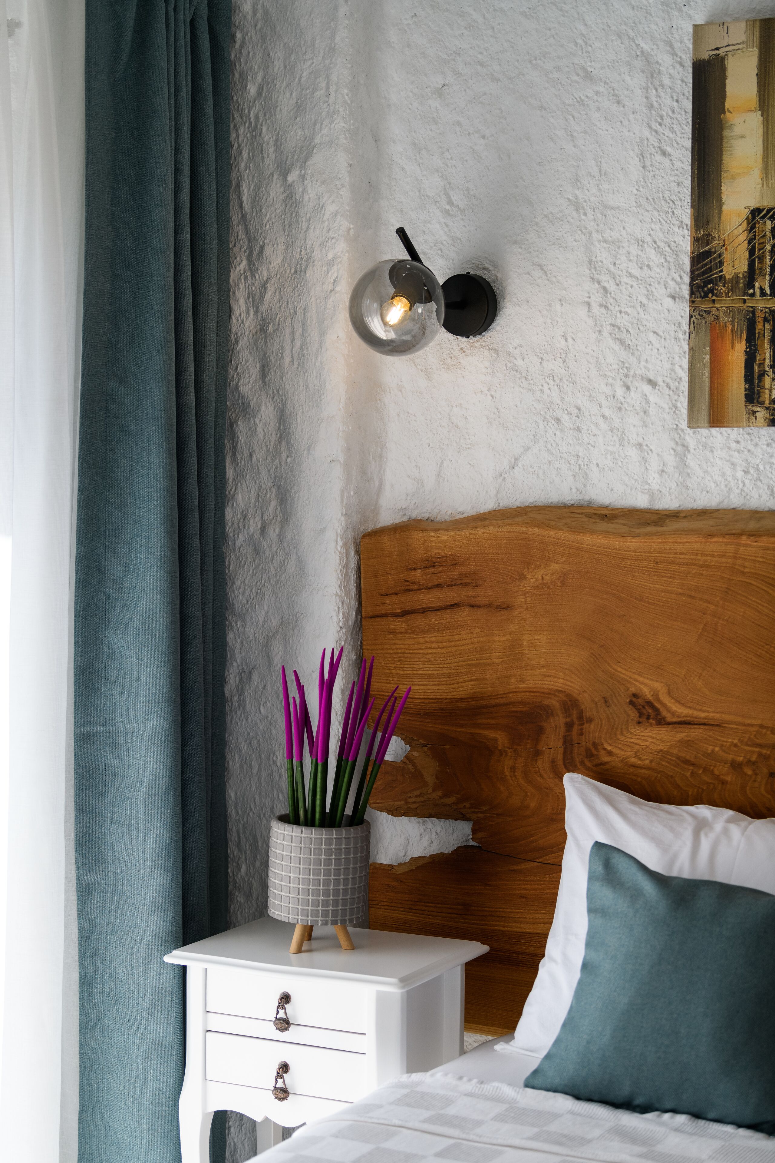 A bedroom with a wooden headboard, a bedside table with drawers, a wall-mounted light fixture, and a window with curtains.
