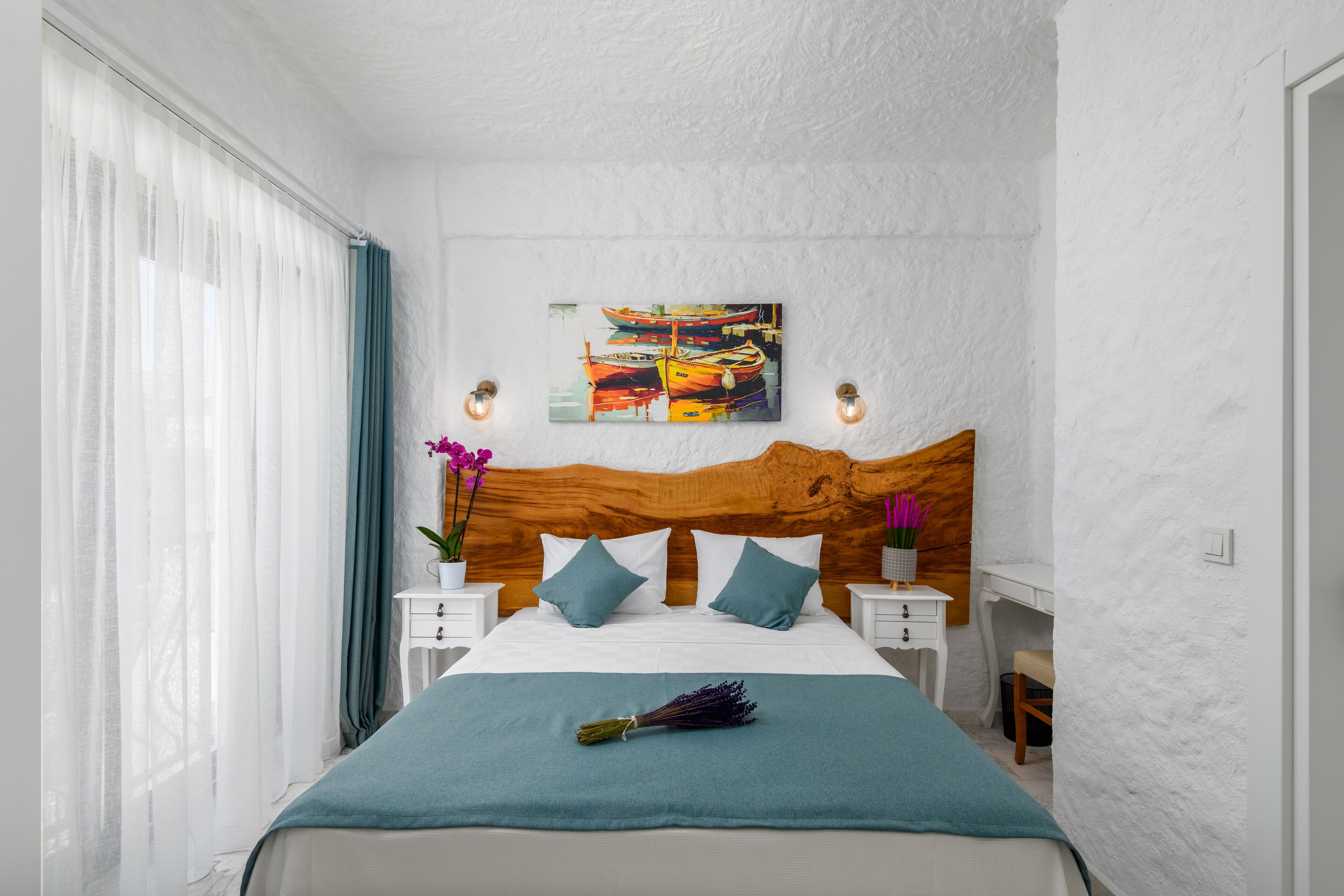 A bedroom with a wooden headboard, white walls, a bed with a teal bedspread, two bedside tables with lamps, and a painting above the bed.