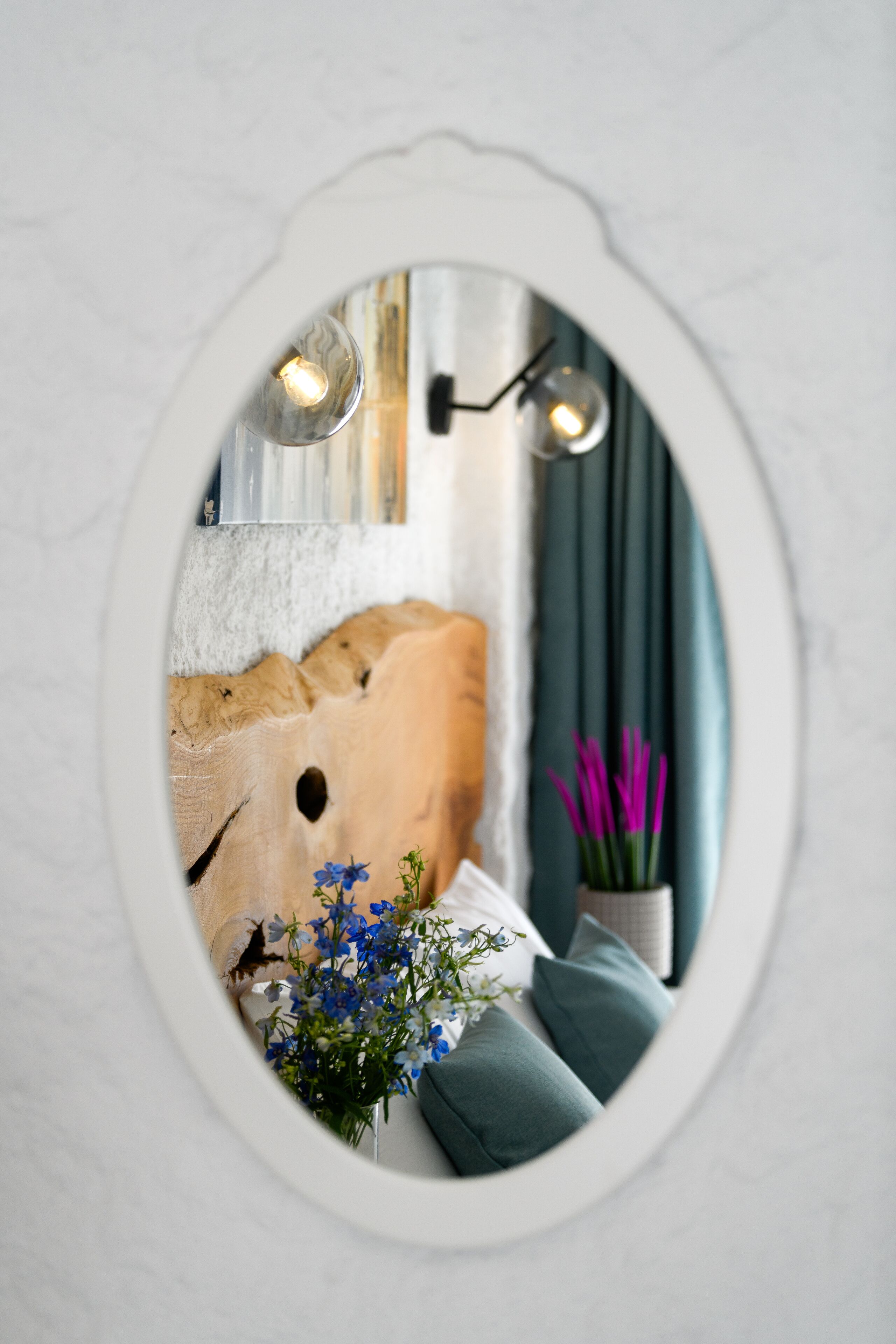 A bedroom with a large mirror, a bed with pillows, a vase with flowers, and a wall-mounted light fixture.