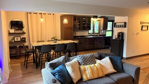 Uma sala moderna com sofá cinza, área de jantar com mesa e cadeiras, e cozinha com armários de madeira e uma geladeira.