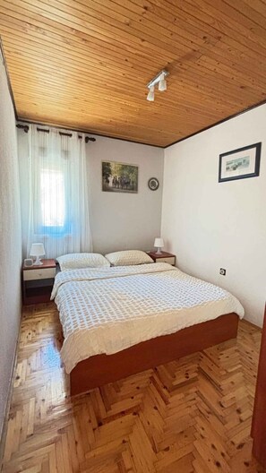 A bedroom with a wooden ceiling, a bed with a white bedspread, a nightstand with a lamp, and a window with curtains.