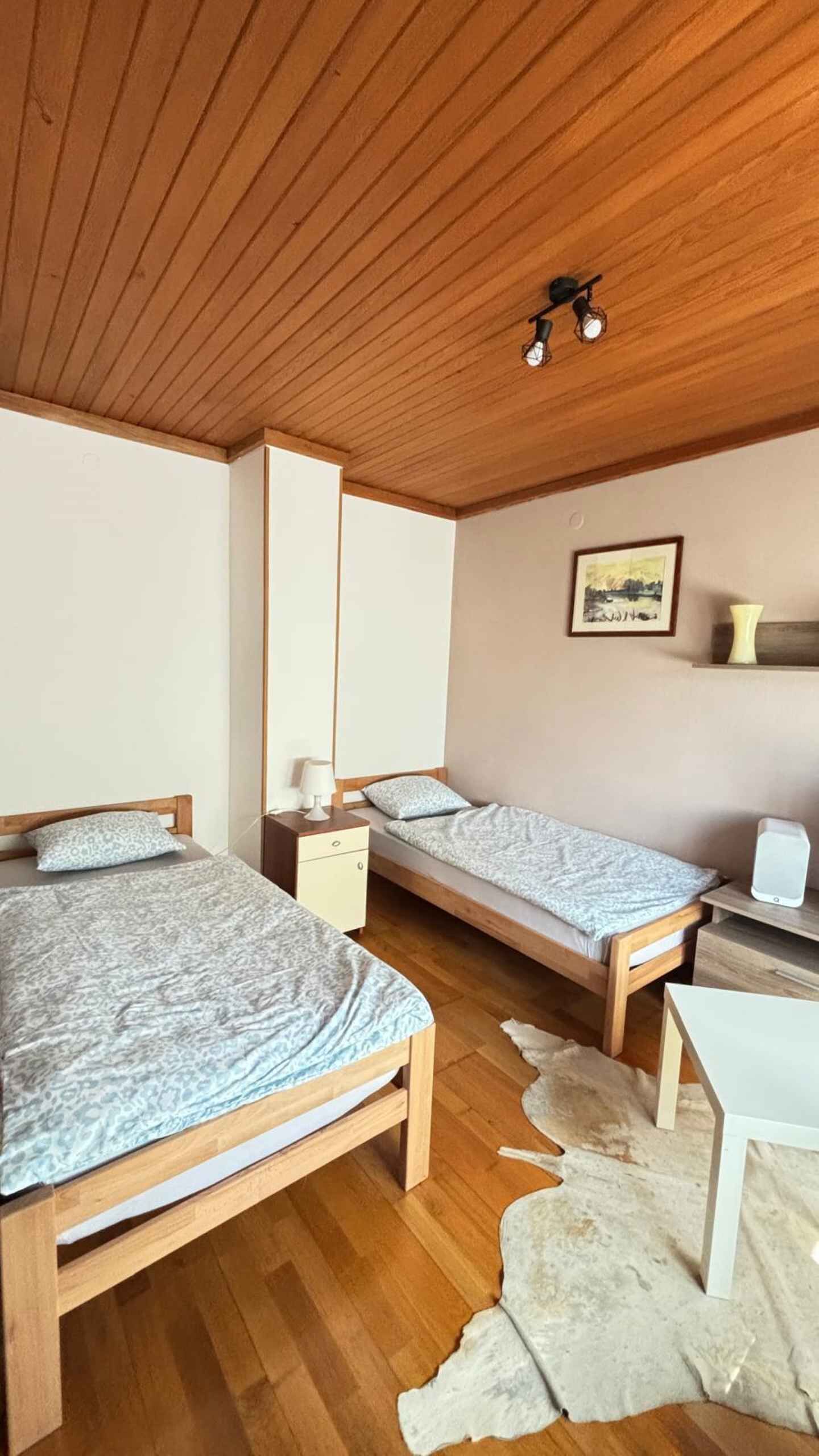 Ett rum med två sängar i trä, ett vitt skrivbord, en liten vit stol, en vit matta och en taklampa.