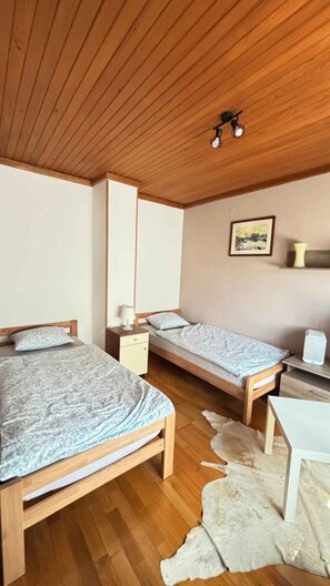 Una habitación con dos camas de madera, un escritorio blanco, una pequeña silla blanca, una alfombra blanca y una lámpara en el techo.