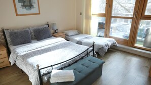 Une chambre avec deux lits, un banc, une table de chevet, une lampe et une fenêtre donnant sur l’extérieur.
