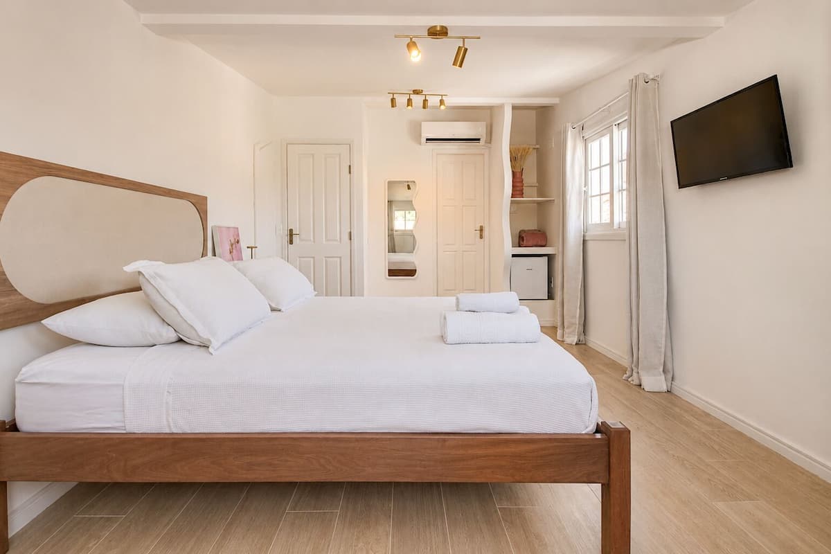 Une chambre avec un lit en bois, du linge de lit blanc, un téléviseur fixé au mur et une salle de bain visible à travers une porte ouverte.