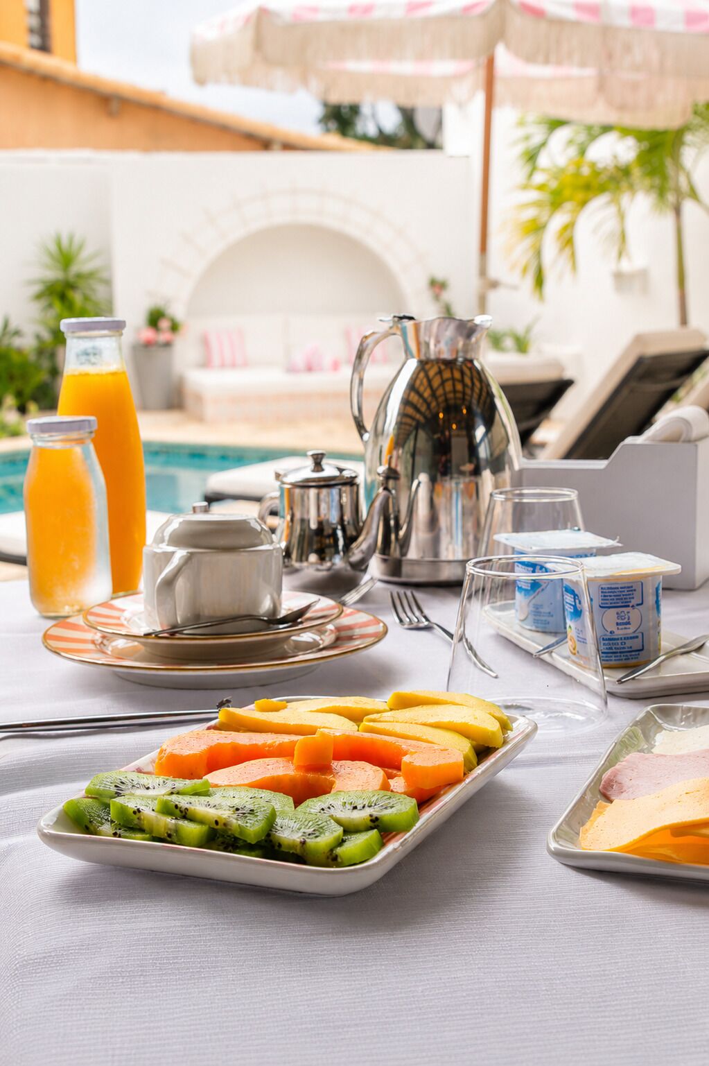A breakfast setup with a fruit platter, a pitcher, a teapot, and a glass of orange juice.