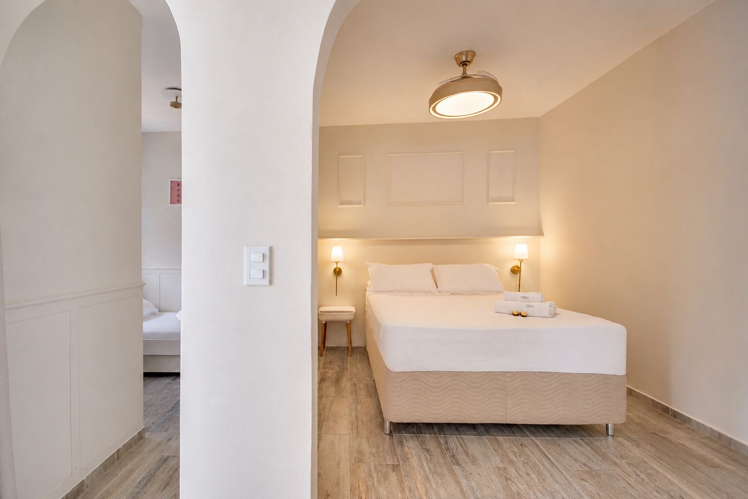 A hotel room with a bed, bedside lamps, a ceiling fan, and a view through an archway to another room.