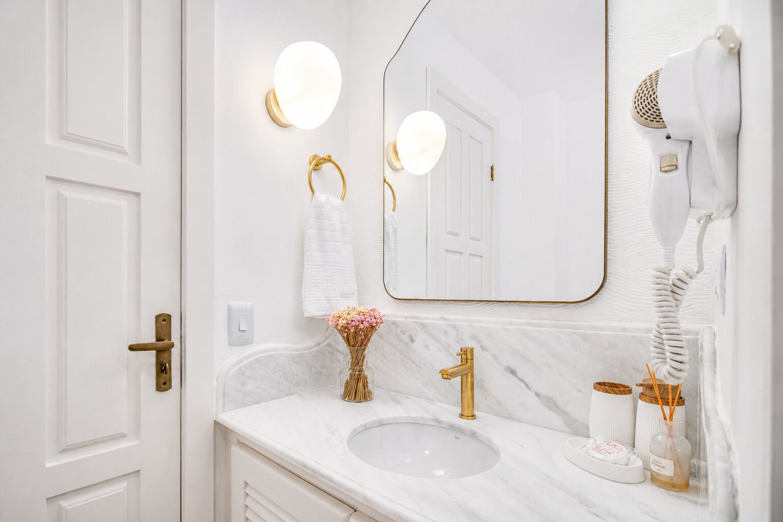 A bathroom with a white marble sink, gold fixtures, a hairdryer, and a vase with flowers.