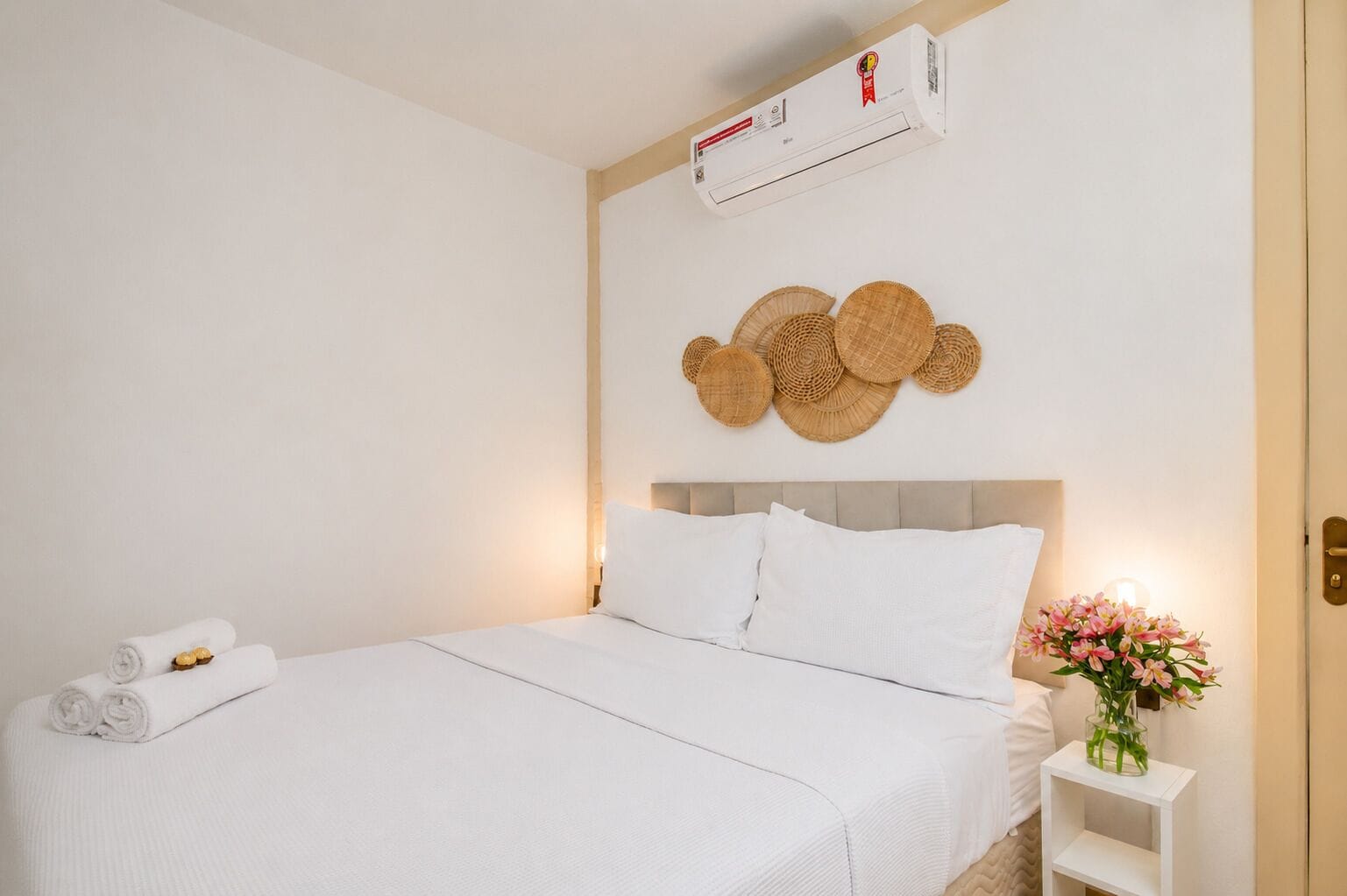 A neatly made bed with white linens, a wall-mounted air conditioner, and a decorative wall hanging.