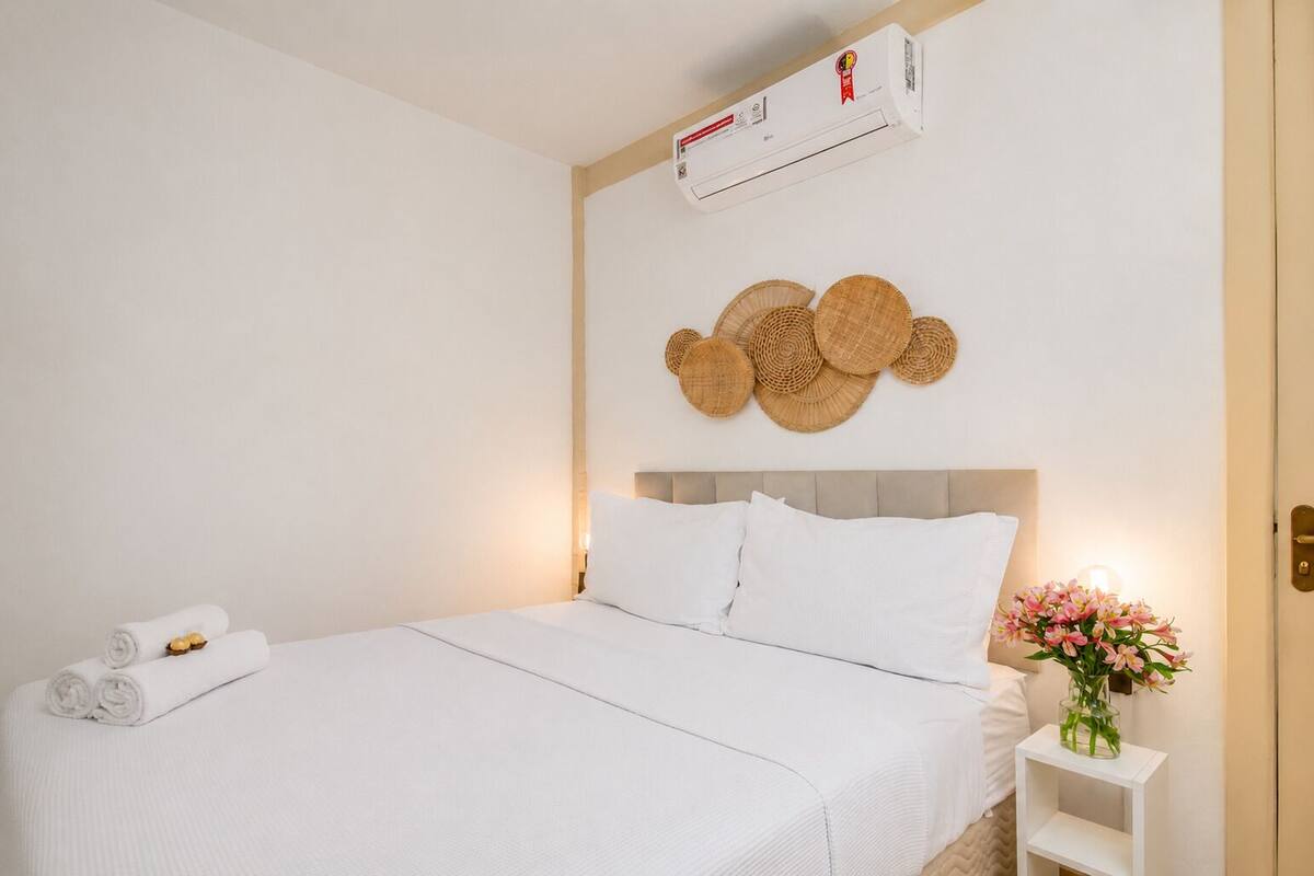 Une chambre d’hôtel avec un lit, un climatiseur fixé au mur, une décoration murale, une table de chevet avec un vase de fleurs et une serviette enroulée sur le sol.