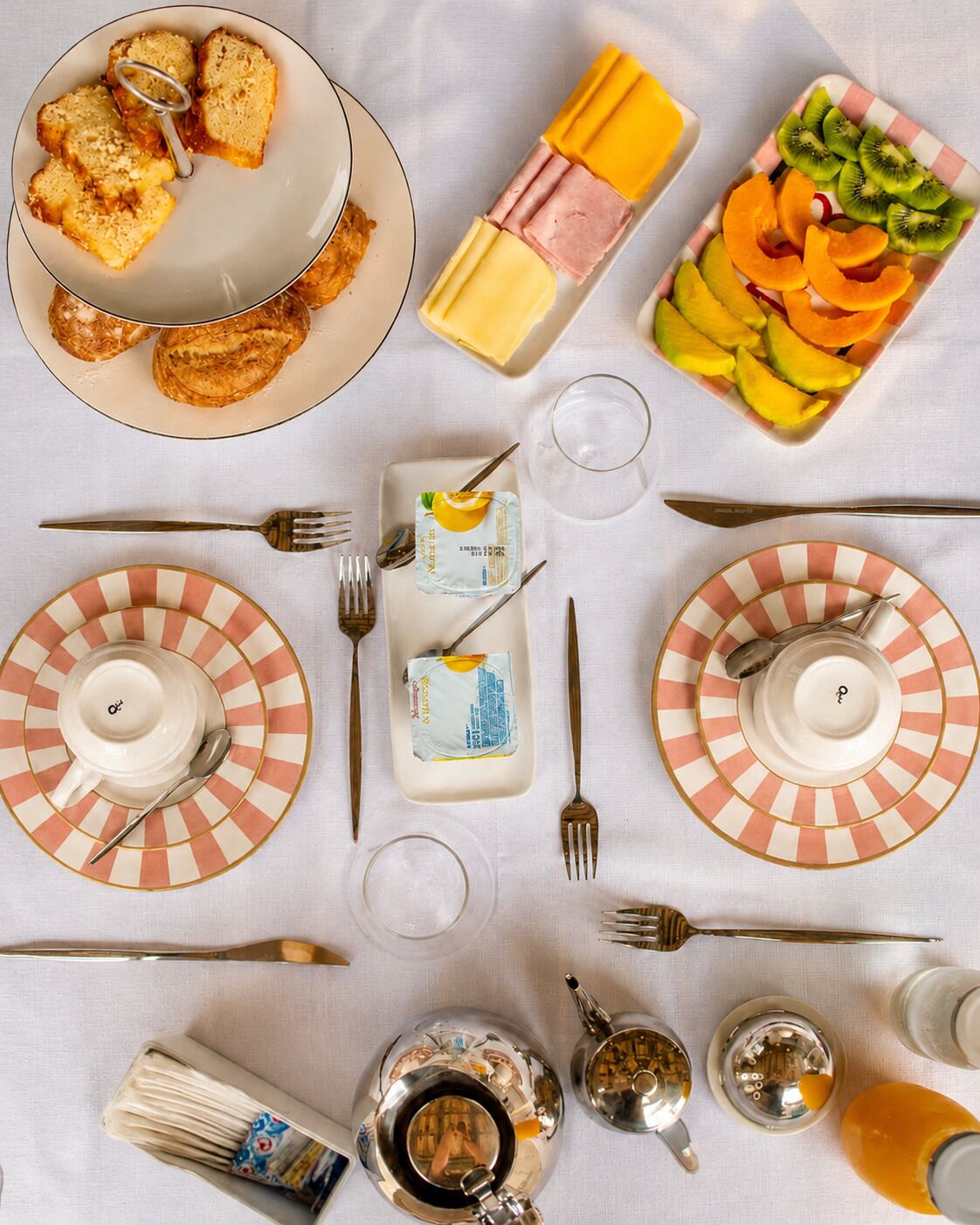 A breakfast setup with toast, cheese, ham, and fruit on plates, along with cutlery and a teapot.