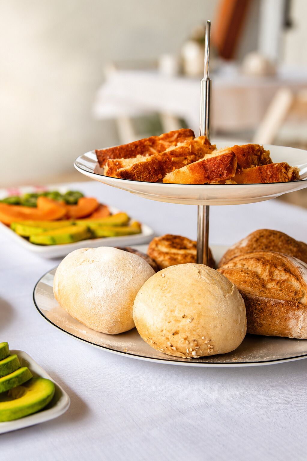 A three-tiered serving tray with bread and pastries, a plate with bread rolls, and a plate with sliced avocado.