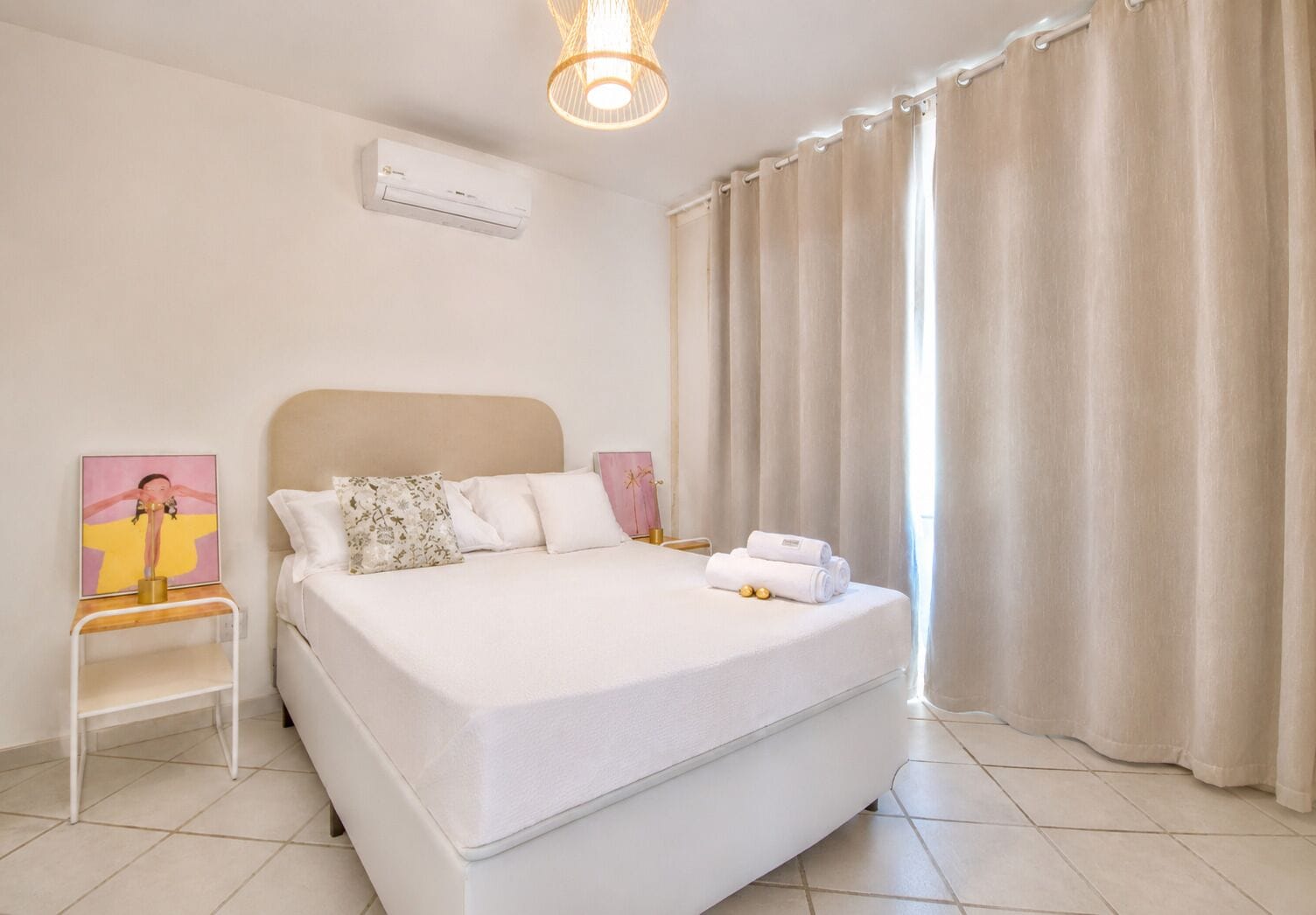 A neatly made bed with white linens, a bedside table with a lamp, and a wall-mounted air conditioner.