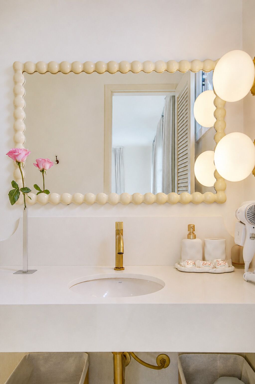 A bathroom vanity with a large mirror, a gold faucet, a vase with pink roses, and a hairdryer.