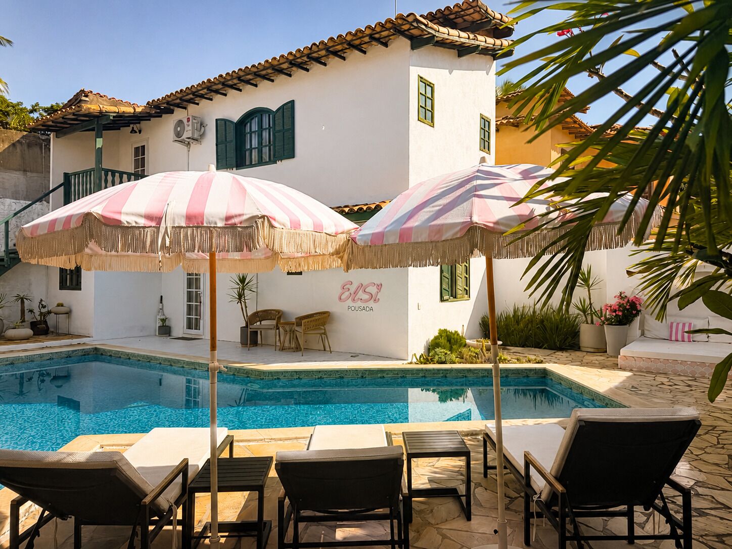 A pool area with lounge chairs and striped umbrellas, a white building with green shutters, and the text 'Elsi Pousada' on the wall.