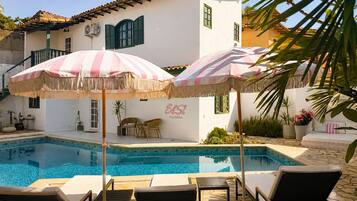 A pool area with lounge chairs and striped umbrellas, a white building with green shutters, and the text 'Elsi Pousada' on the wall.